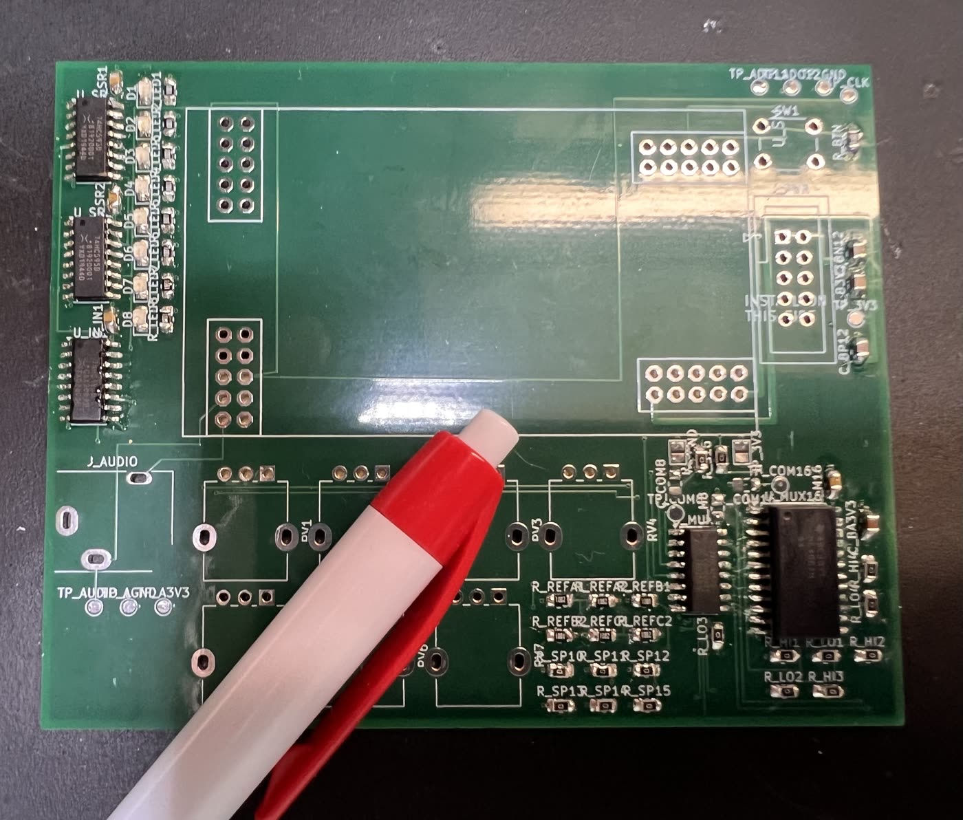 The whole 100 × 80 mm board lying on a dark mat with a red and white pen across it. Down the left edge three black SOIC chips are soldered in place, a row of small resistors above them and the LED pads between still empty. The middle of the board is the large empty outline the Daisy Patch Submodule seats into. At the bottom right the two mux chips are fitted and surrounded by dozens of tiny resistors, some places filled and some still bare gold. The potentiometer footprints and the audio jack along the bottom left have not been touched.