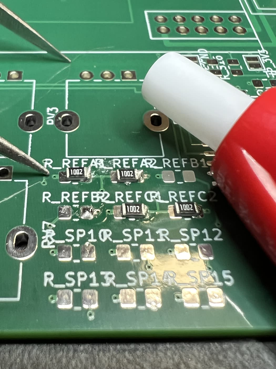 A close-up of the reference-divider corner. Four black 0805 resistors marked 1002 sit on their pads in two rows, under silkscreen names that overlap into each other: R_REFA1, R_REFA2, R_REFB1, R_REFB2, R_REFC1, R_REFC2. The pads of R_REFB2 carry a bright, over-large bead of solder that covers both ends of the part. A pair of tweezers comes in from the left and the tip of a red pen from the right. Below them the R_SP pads are still bare gold.