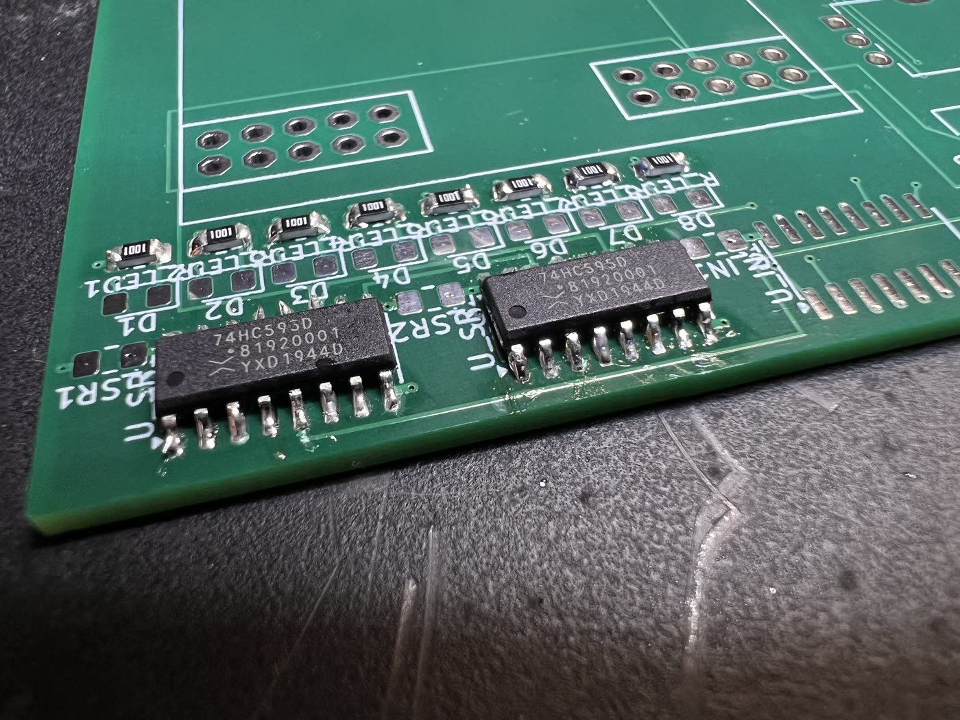Two 74HC595D shift registers soldered onto their footprints at the corner of the board, seen from a low angle so the light catches every leg. Above them a row of 0805 resistors marked 1001 sits under the names R_LED1 to R_LED8, with the LED pads between them still empty.