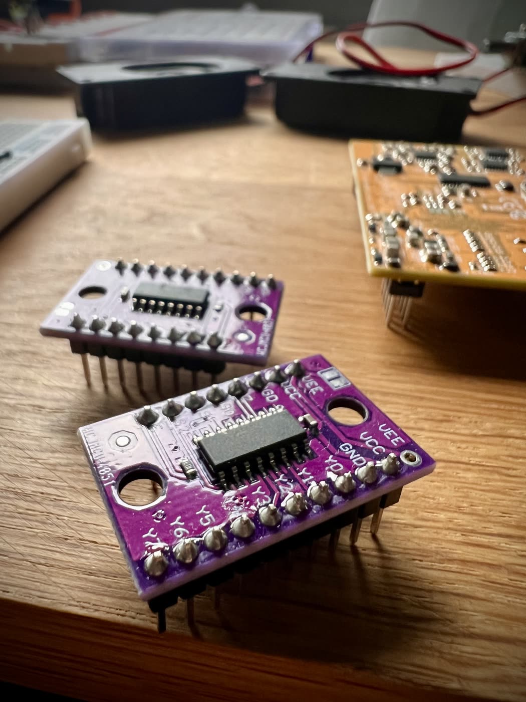 Two small purple breakout boards on a wooden desk, each carrying a single wide-body chip and a row of header pins soldered through. The nearer one is turned so its silkscreen reads: Y0 to Y7 down one side, GND, VCC and VEE at the corner, the select lines along the top. Behind them sit an orange circuit board, a couple of black round components and a bare breadboard, all out of focus.